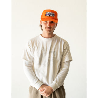 WELCOME SKATEBOARDS. MODEL WEARING: ORANGE TRUCKER HAT WITH MESH BACK FOAM FRONT. SNAP CLASP. WELCOME LOGO WITH PURPLE THORNS ACROSS LOGO ON FRONT. ORANGE BRAID ALONG BILL.