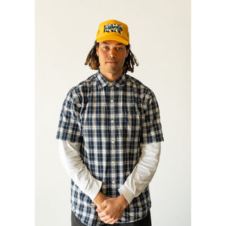 MODEL WEARING WELCOME SKATEBOARDS. GOLD TRUCKER HAT WITH MESH BACK FOAM FRONT. SNAP CLASP. WELCOME LOGO WITH GREEN THORNS ACROSS LOGO ON FRONT. GOLD BRAID ALONG BILL.