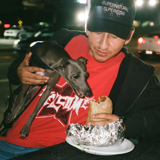 Skater in a Supernatural Superstar trucker hat and red Welcome tee, holding a dog and a burrito at night