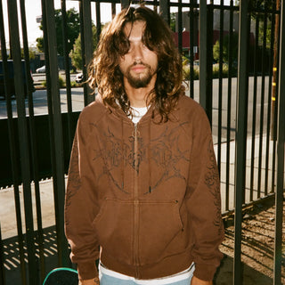 Skater in a brown embroidered zip hoodie standing at a black iron fence